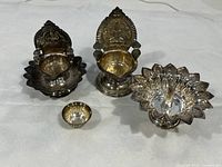 Three silver metallic diya candlestick holders and a small rounded bowl displayed on white surface.
