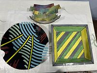 Four multicolor signed ceramic decorative pieces: large round plate, square plate, and two small bowls with gradient colors placed on a white cloth.