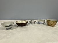 Five small decorative bowls arranged in a row on a white cloth against a gray background.