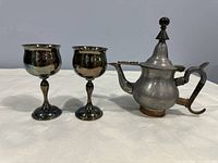 Front view of teapot with lid closed and two small silver-plated goblets standing beside it, showing polished exterior with patina.
