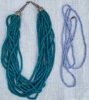 Photo of two beaded necklaces on a white textured background, showing the multi-strand turquoise necklace and the single strand light purple and white striped necklace.