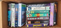 Box with 9 assorted VHS tapes showing titles and cases stacked upright