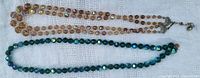 Two necklaces laid flat showing their full length: one light brown faceted bead necklace with iridescent highlights and one dark green faceted bead necklace with iridescent highlights.