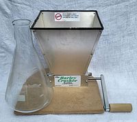 Side view of The Barley Crusher Malt Mill showing grain hopper, wooden handle crank, wood base and adjacent 2000ml glass beaker.