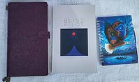 Front view of three notebooks: leather bound purple notebook, 'Blind Pilot' book, and spiral bird cover notebook.