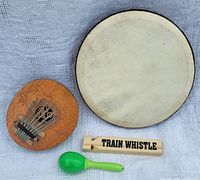All four musical instruments displayed on light fabric background. Includes hand drum, kalimba, green shaker, and wooden train whistle.