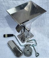 Front view of manual stainless steel tomato press with crank handle and clamp attachment, showing hopper and press components spread out.