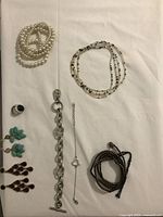 Photo of assorted jewelry laid out on white cloth showing pearl necklace, silver bracelets, various earrings, and rings.