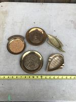 Top-down photo of five brass decorative items displayed on a white surface with measuring tape showing approximate sizes, highlighting the shape and design of each piece.