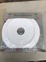 Full view of unopened ceiling medallion packaged in clear plastic with a label header showing 'Decora Ornamental set of 1 decoration' with embossed decorative pattern visible.