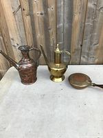 Three items on a table against a wooden fence: copper pot on left, brass pot in center, and brass crumb catcher pan on right.