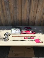 Photo of the lot items on a table against wooden fence; showing dog bowls, harness, collars, leash, and potty pads.