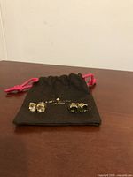 Two sets of stud earrings on a dark wooden surface with a black Kate Spade dust bag with pink drawstrings behind them.