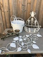 White metal hanging wall planter, decorative white metal scroll leaf panel, three white metal bucket style planters with wire handles, and green metal 3-candle holder on concrete surface with wooden fence background.
