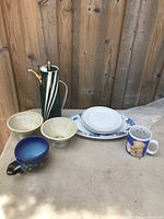Overview of kitchen ceramics set on table in front of wooden fence, shows all items including jug, bowls, plates, and mugs.