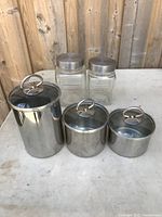 Front view of the five kitchen storage containers showing 3 stainless steel canisters with clear lids and 2 glass jars with metal screw lids on a wooden background.