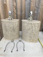 Front view of two beige MCM style table lamps with perforated cylindrical bases and metal harp frames for lamp shades on a light surface in front of a wood fence.