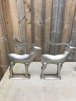 Two silver-tone metal reindeer standing side by side on a gray table against wood panel background. Photo shows full front and side profile of the decorations.