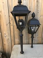 Photo showing one large and one small black cast iron outdoor lamp with a classical lantern design and clear glass panels on a wood background.