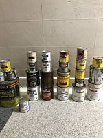 Full display of 17 assorted paint, wood stain, and finish cans including larger and smaller sizes grouped in front of a beige wall.