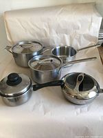 Five stainless steel cookware pieces on white background including pots, saucepans, and lids with black and metal handles.