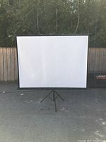 Front view of the brand new 60-inch projection screen set up on black tripod stand outdoors with fence and trees in background.