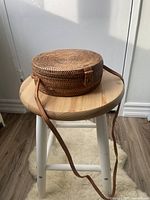 Side view of round woven rattan bag resting on a wooden stool, showing the circular shape and leather strap.