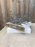 Two white floating shelves stacked with a silver metal decorative bowl on top showing leaf design and bottom shelf mounting hardware.