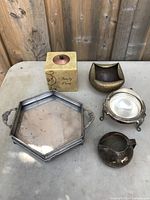 Photo of silver-plated hexagonal tray with handles, cube candle holder, brass bowl, silver-plated bowl with feet, and small pitcher on a table against wood background