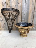 Gold-colored decorative vase with black interior next to black metal wall ornament with intricate scroll designs, shown on plain surface against wood background.
