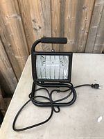 Front and side view of a black metal electric work lamp with protective grill and foam handle grip on a white surface.
