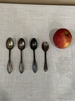 Photo showing three silver spoons with leaf-patterned handles and one smaller silver plated spoon placed on fabric with an apple for size reference.
