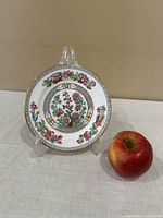 Decorative plate with floral design including pink, blue, and green flowers and a geometric border, propped on a stand next to an apple for scale
