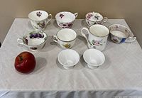 Nine assorted decorative teacups arranged with an apple on a tablecloth, showing front and side views.
