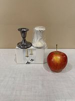 Two candle holders and one candle with an apple for size reference, one holder is metal and the other candle holder is wrapped in plastic.