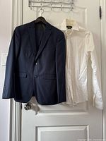 Dark navy suit jacket and white shirt hanging on door. Suit is slim fit, shirt shows wrinkles and needs cleaning.