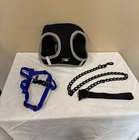 Black mesh dog harness with silver trim, blue adjustable nylon harness, and heavy black dog chain with attached strap on a white cloth surface.