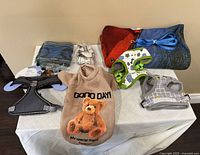 Top view of eight assorted dog supplies arranged on a white surface including harnesses, a hoodie, dog coats, and leashes