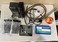 Fish tank supplies including the main filter unit, tubing, and mesh filter bags arranged on a table with an apple for scale.