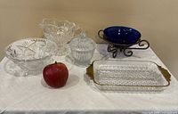 Five glass items on white surface with apple for scale, including clear pressed glass bowls with starburst patterns, covered dish, cobalt blue glass bowl on stand, and rectangular tray with brass handles.