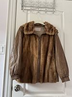 Front view of the jacket showing brown fur front panels and brown leather sleeves with elastic cuffs, hanging on door.