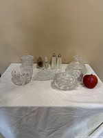 Collection of assorted glass items including bowls, salt and pepper shakers, decanter, and dishes arranged on a white cloth with an apple for scale.