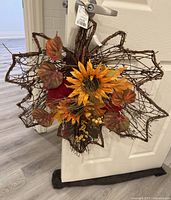 Photo of a fall-themed door decor wreath hanging on a door, showing the woven twig base decorated with artificial sunflowers, autumn leaves, and berries, with tag attached indicating it is new.