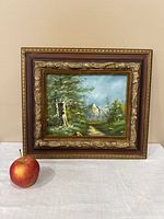 Small square landscape painting on plywood with a detailed forest path and mountain scene framed in a carved gold and brown wooden frame, photographed against a neutral wall with an apple placed beside for size reference.