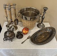 Overview of silver plated ice bucket with woven outer basket, supported by two handles, with set of three serving utensils on a stand, two candle holders, an oval tray, a two-prong fork, and a covered sugar bowl on a white tablecloth.