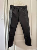 Front view of black leather pants hanging on a hanger against a white door. Shows slim fit design, front pockets, and attachment details.