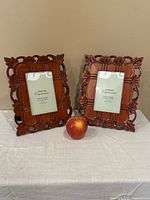Two matching carved wooden picture frames side by side with an apple for scale, showing decorative floral edges and a 3x5 inch photo placeholder inside each.