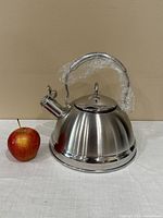 Full side view of the tea kettle next to an apple for scale, showing the metallic finish, handle with plastic wrap, and spout with lever.