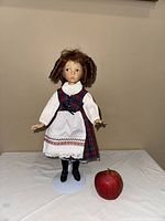 Front view of porcelain doll with curly brown hair wearing traditional red plaid dress next to an apple for size reference.