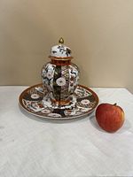 Full set of Saji fine china including vase with lid placed on matching plate alongside a red apple for scale.
